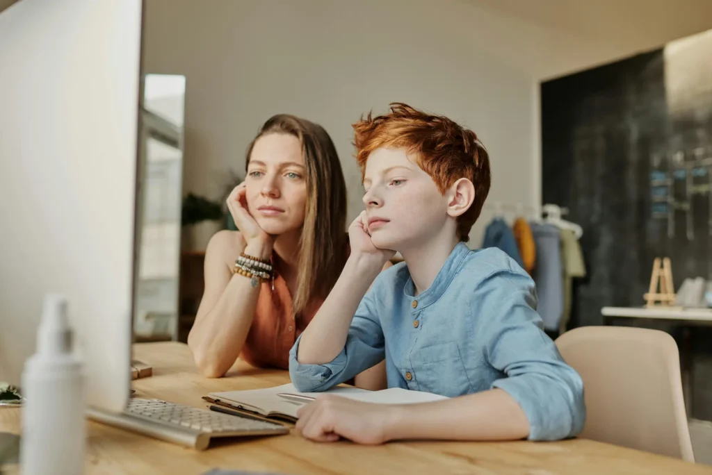 A woman and a boy are seated at a desk, both focused on a computer screen in front of them.