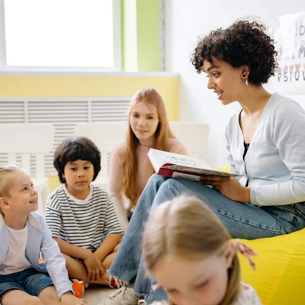 A woman engagingly reads a story to a group of attentive children in a bright classroom setting.